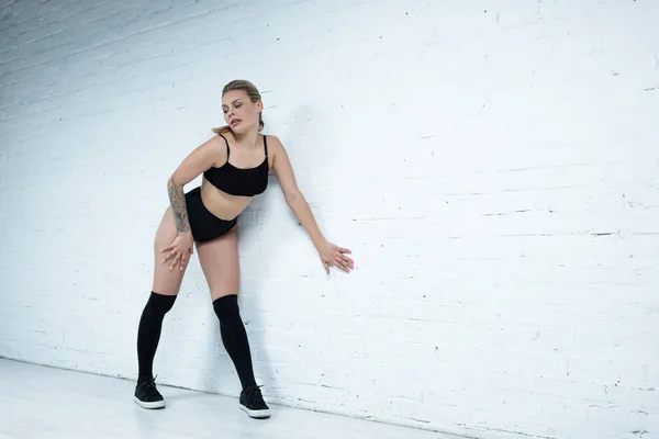 Tattooed sexy blonde girl in black outfit twerking near wall in studio — Stock Photo