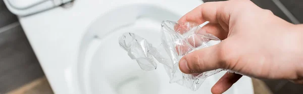 Vista cortada do homem jogando crumpled garrafa de plástico em vaso sanitário em banheiro moderno com azulejo cinza, tiro panorâmico — Fotografia de Stock