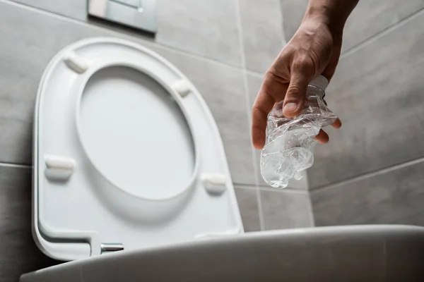 Обрезанный вид человека, бросающего мятую пластиковую бутылку в унитаз в современном туалете с серой плиткой — стоковое фото