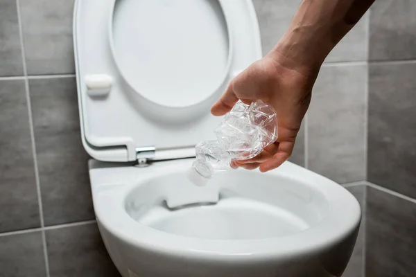 Vista cortada do homem jogando crumpled garrafa de plástico em vaso sanitário no banheiro moderno com azulejo cinza — Fotografia de Stock