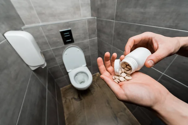 Vista cortada do homem segurando pílulas perto de vaso sanitário cerâmico limpo no banheiro moderno com azulejo cinza — Fotografia de Stock