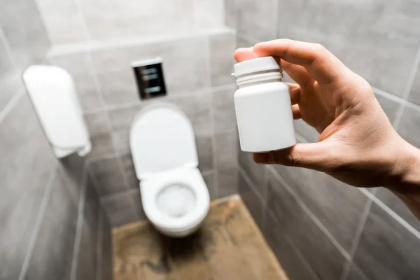 Vista cortada do homem segurando recipiente com pílulas perto de vaso sanitário cerâmico limpo no banheiro moderno com azulejo cinza — Fotografia de Stock