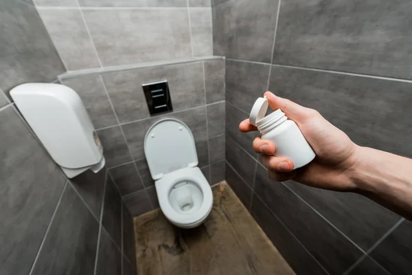 Abgeschnittene Ansicht eines Mannes, der Behälter mit Pillen in der Nähe von Keramik aufbewahrt, saubere Toilettenschüssel in einer modernen Toilette mit grauer Fliese — Stockfoto