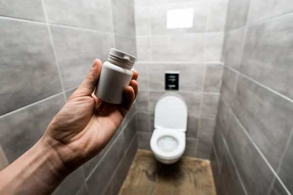 Обрізаний вид людини, що тримає тару з таблетками біля керамічного чистого унітазу в сучасному туалеті з сірою плиткою — стокове фото