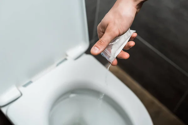 Обрезанный вид человека, выливающего кокаин в унитаз в современном туалете с серой плиткой — стоковое фото