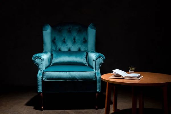 Elegante poltrona blu velluto vicino a tavolo in legno con libro isolato su nero — Foto stock