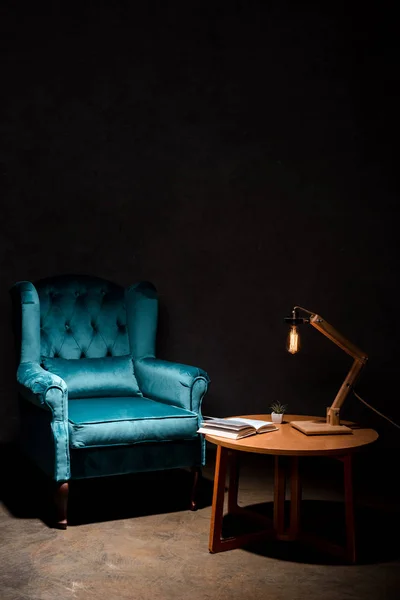 Élégant fauteuil en velours bleu avec oreiller près de la table en bois et lampe isolée sur noir — Photo de stock