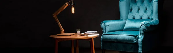 Elegant velour blue armchair with pillow near wooden table and lamp isolated on black, panoramic shot — Stock Photo