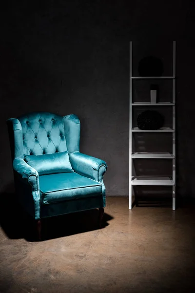Elegant velour blue armchair with pillow near rack in dark room — Stock Photo
