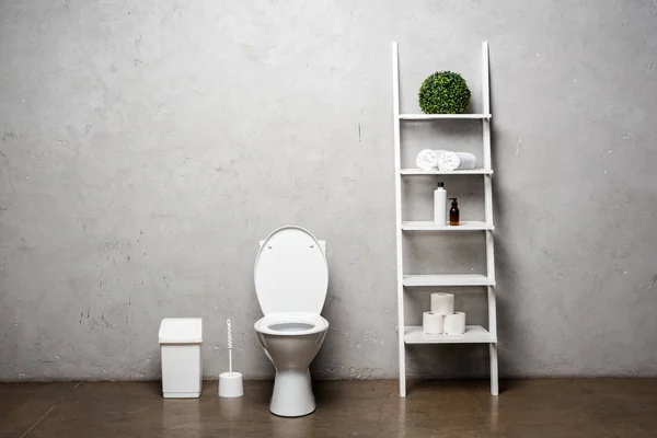 Intérieur de la salle de bain moderne avec cuvette de toilette près de rack avec cosmétiques, serviettes, papier toilette, poubelle et brosse de toilette — Photo de stock