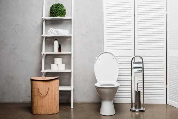 Interior of modern bathroom with toilet bowl near rack with cosmetics, towels, toilet paper, laundry basket and toilet brush — Stock Photo