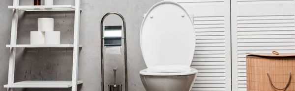 Interno del moderno bagno con wc vicino a rack con asciugamani, carta igienica, cestino della lavanderia e spazzola da toilette, colpo panoramico — Foto stock