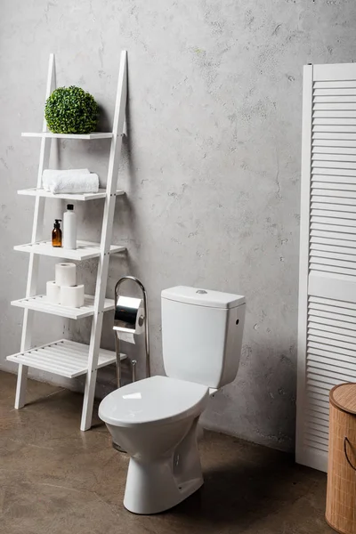 Intérieur de la salle de bain moderne avec cuvette de toilette près de rack avec cosmétiques, serviettes, papier toilette, panier à linge et brosse de toilette — Photo de stock