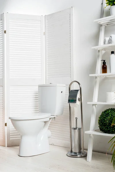 Interior of white modern bathroom with toilet bowl near folding screen, rack and plants — Stock Photo