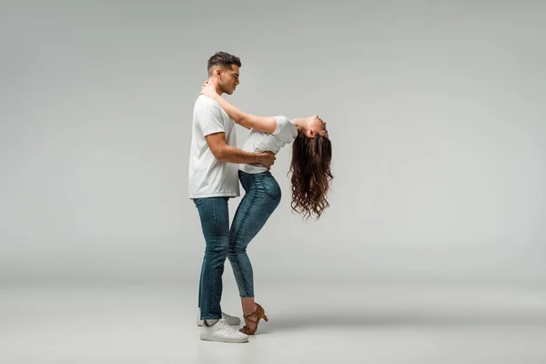 Vista lateral de dançarinos em camisetas e jeans bachata dançando em fundo cinza — Fotografia de Stock