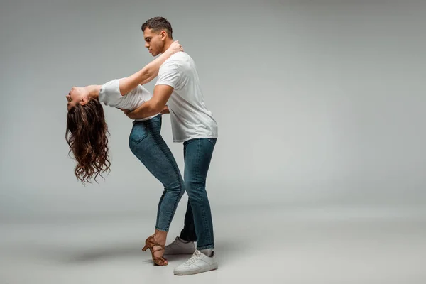 Vista lateral de bailarinas en camisetas y jeans bailando bachata sobre fondo gris - foto de stock