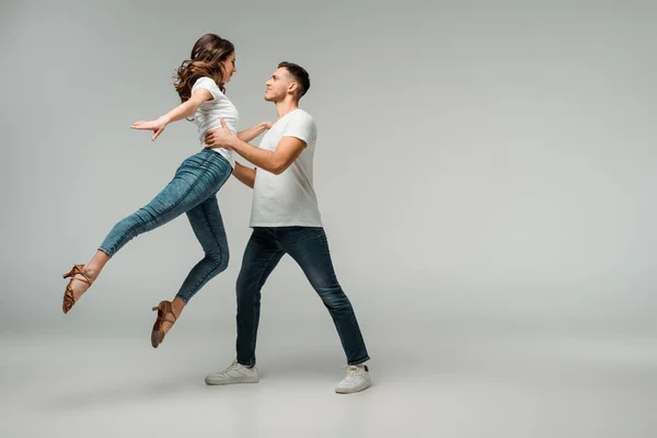 Vue latérale de danseurs souriants en t-shirts et jeans dansant bachata sur fond gris — Photo de stock