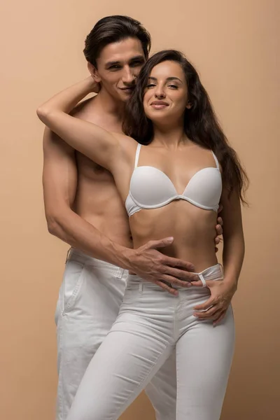 Uomo senza maglietta che abbraccia donna felice in reggiseno bianco, isolato sul beige — Foto stock