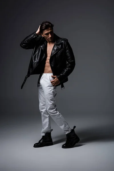 Мужчина без рубашки в белых джинсах и черной кожаной куртке, на сером — стоковое фото