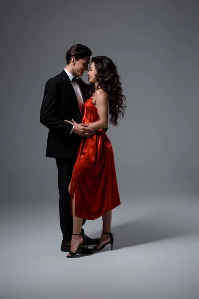 Pareja romántica en traje y vestido de seda roja abrazándose y mirándose en gris - foto de stock