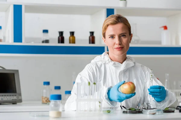 Biologist looking at camera and holding whole apple and test tube in lab — Stock Photo