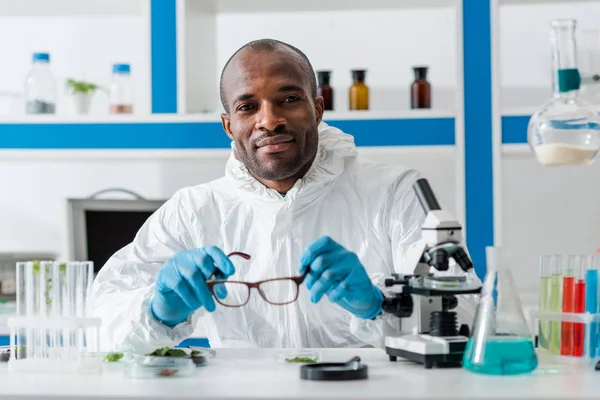 Smiling african american biologist holding glasses and looking at camera — Stock Photo