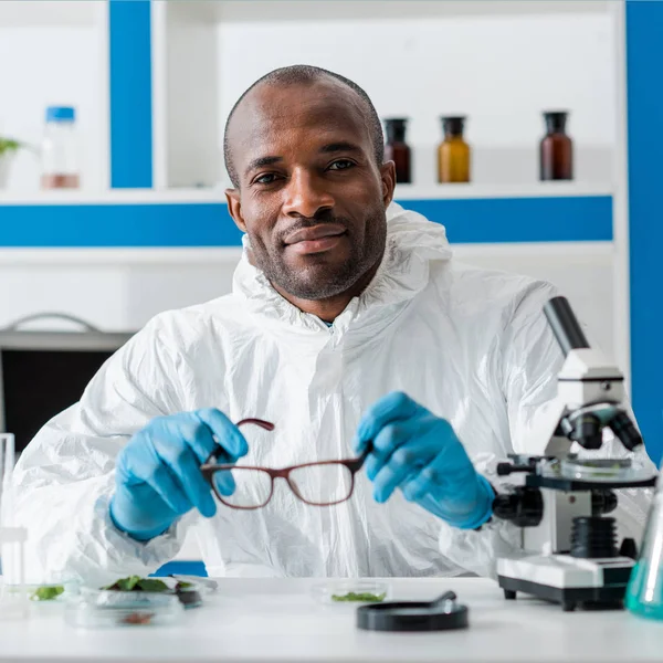 Smiling african american biologist holding glasses and looking at camera — Stock Photo