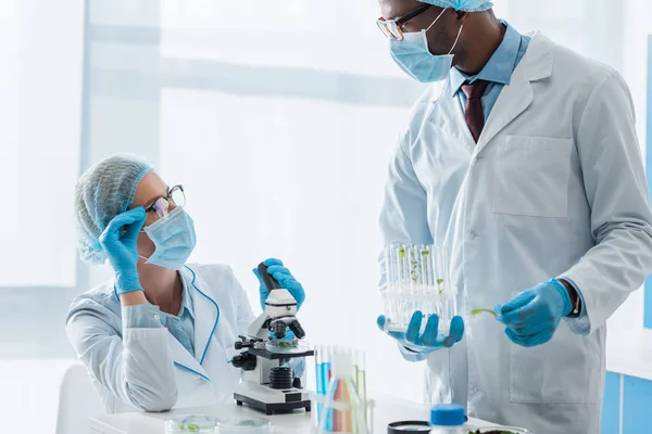 Biólogos multiculturales con batas blancas y máscaras médicas hablando en laboratorio - foto de stock