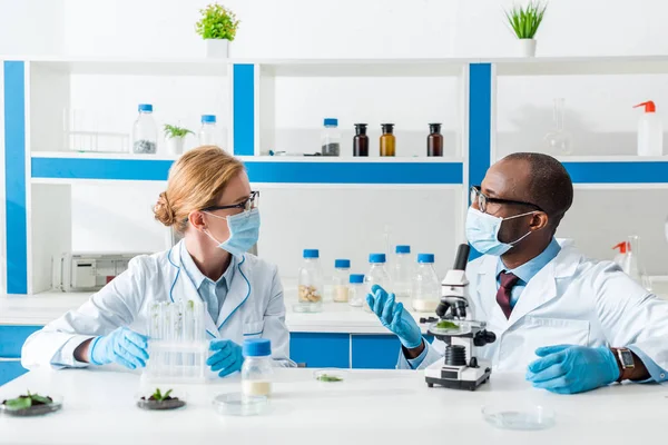 Biólogos multiculturais em casacos brancos e máscaras médicas falando em laboratório — Fotografia de Stock