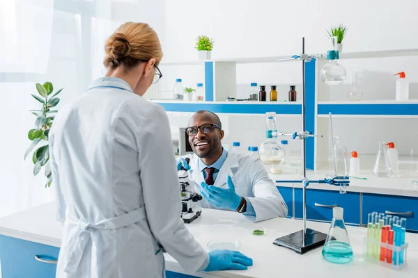 Souriant biologiste afro-américain assis à table et regardant son collègue — Photo de stock