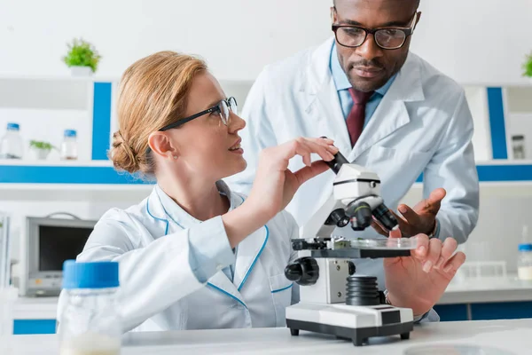 Biólogos multiculturais em óculos olhando microscópio em laboratório — Fotografia de Stock