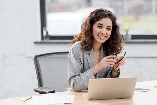 Atractivo y sonriente gerente de cuenta sentado en la mesa y sosteniendo los vasos — Stock Photo