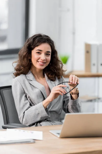 Atractivo y sonriente gerente de cuenta sentado en la mesa y sosteniendo los vasos - foto de stock