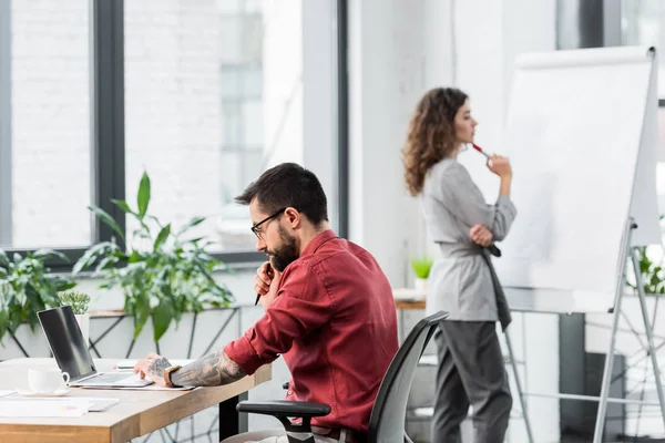 Enfoque selectivo del administrador de cuentas utilizando el ordenador portátil y su colega de pie cerca de flipchart - foto de stock