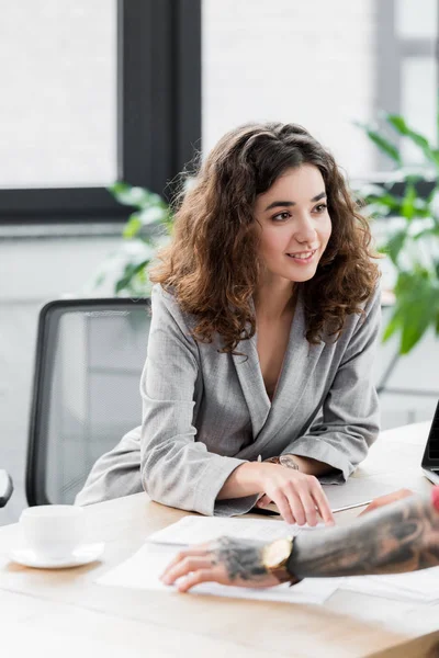 Sonriente gerente de cuenta sentado en la mesa y mirando a su colega - foto de stock