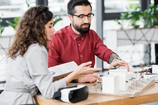 Arquitecto de realidad virtual apuntando con el modelo de mano en casa y hablando con colega - foto de stock