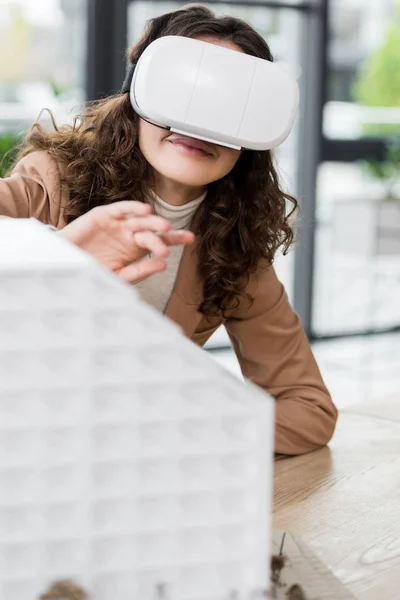 Селективный фокус улыбающегося архитектора виртуальной реальности в гарнитуре виртуальной реальности, смотрящего на модель дома — стоковое фото