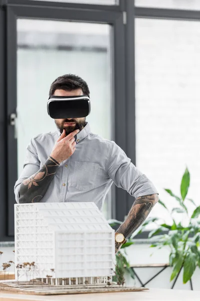 Pensive virtual reality architect in virtual reality headset looking at model of house — Stock Photo