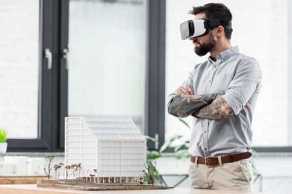 Virtual reality architect with crossed arms in virtual reality headset looking at model of house — Stock Photo