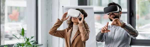 Panoramic shot of smiling virtual reality architects in virtual reality headsets pointing with finger — Stock Photo