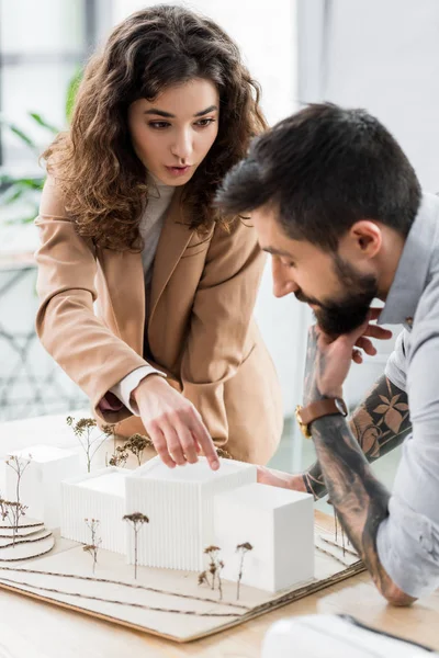 Enfoque selectivo del arquitecto de realidad virtual apuntando con el dedo al modelo de casa y hablando con su colega - foto de stock