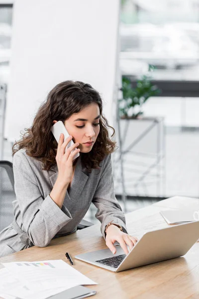 Attractive account manager sitting at table and talking on smartphone in office — Stock Photo