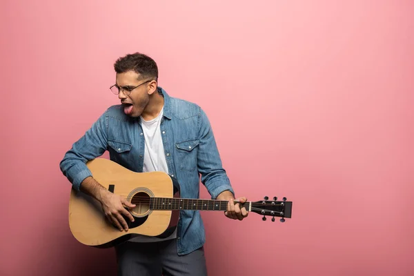 Jovem saindo da língua enquanto tocava guitarra acústica no fundo rosa — Fotografia de Stock