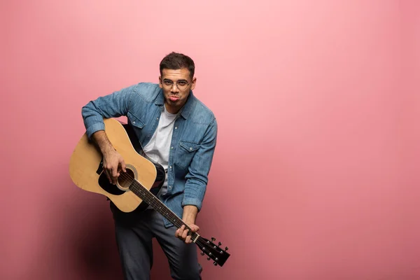 Schöner Mann grimmig, während er auf einer Akustikgitarre auf rosa Hintergrund spielt — Stockfoto