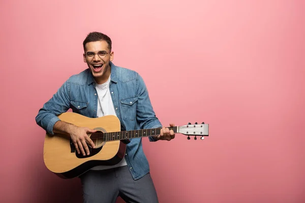 Jovem homem rindo olhando para a câmera enquanto tocava guitarra acústica em fundo rosa — Fotografia de Stock