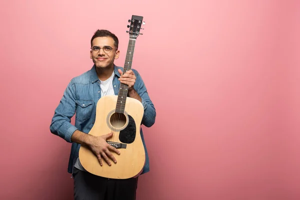 Homem sorrindo para a câmera e segurando guitarra acústica em fundo rosa — Fotografia de Stock