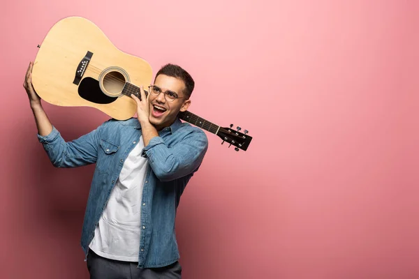 Веселый человек с акустической гитарой, смотрящий в сторону на розовом фоне — стоковое фото