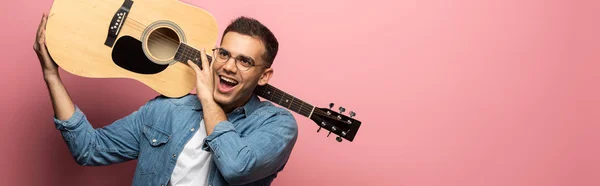 Panoramic shot of excited man with acoustic guitar looking away on pink background — Stock Photo