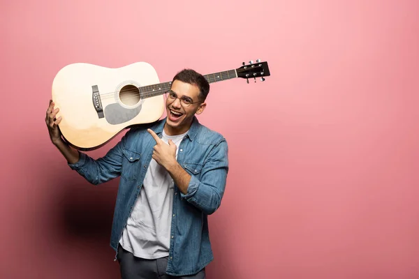 Веселый мужчина смотрит в камеру и указывает на акустическую гитару на розовом фоне — стоковое фото