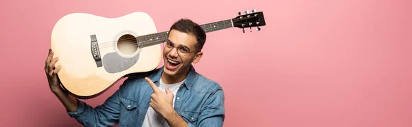 Panoramaaufnahme eines fröhlichen Mannes, der auf eine Akustikgitarre auf rosa Hintergrund zeigt — Stockfoto